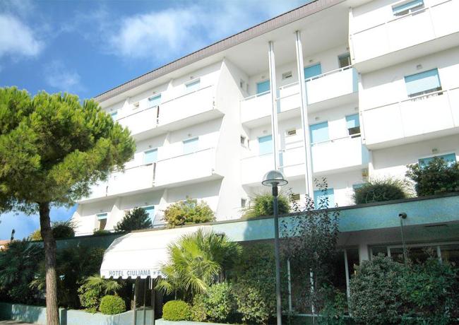 Hotel moderno con balconi, circondato da alberi e vegetazione.