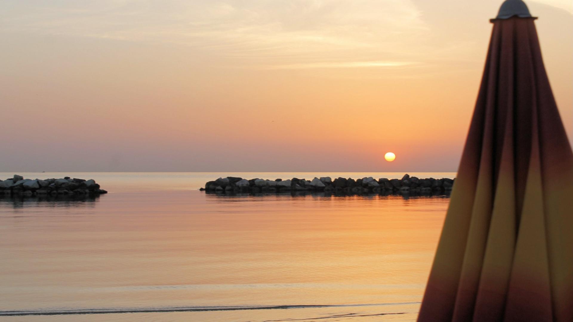 Tramonto sereno sul mare calmo, ombrellone chiuso e cielo arancione riflesso sull'acqua.