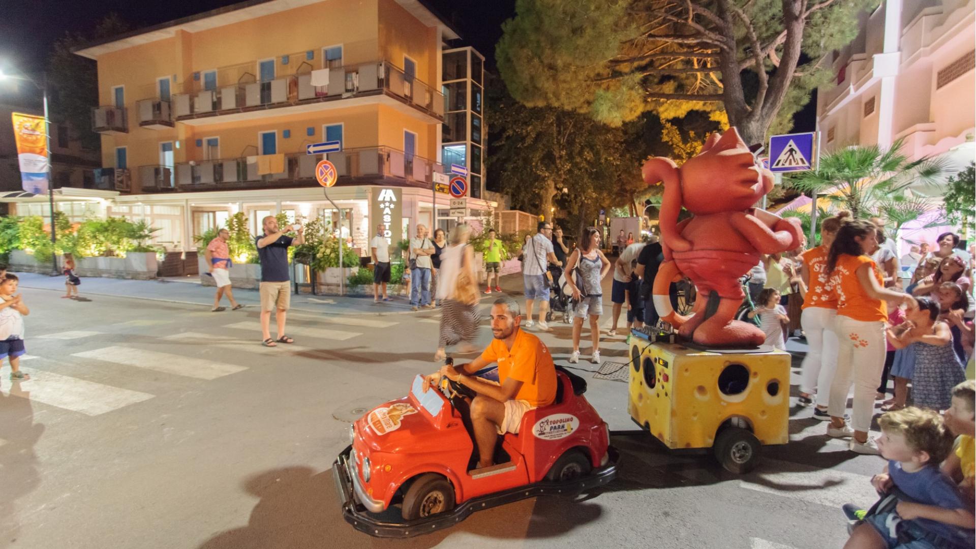 Parata notturna festosa, auto giocattolo rossa e folla sorridente in una strada illuminata.