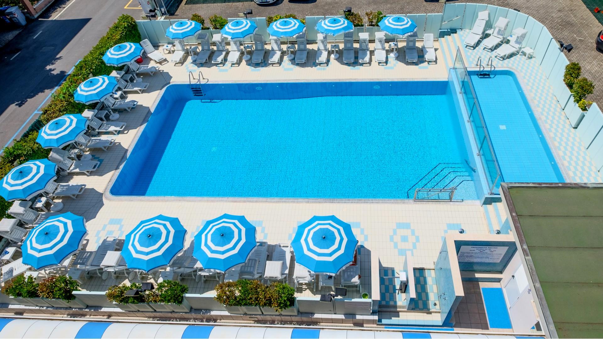 Piscina dall'alto, ombrelloni blu, atmosfera estiva, relax e tranquillità.