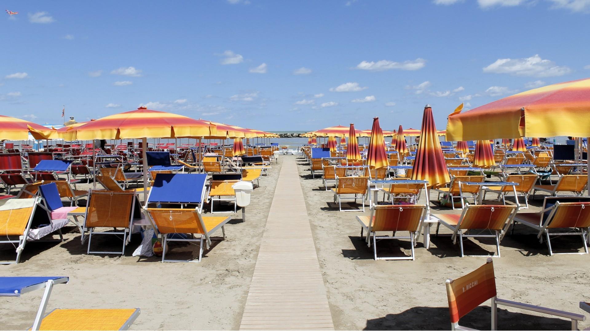 Spiaggia assolata con ombrelloni colorati, sedie a sdraio e cielo azzurro punteggiato di nuvole.