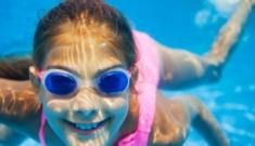 Bambina felice nuota sott'acqua, indossa occhialini blu e costume rosa, luce solare vibrante.