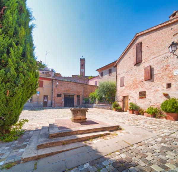 Piazza tranquilla con edificio in mattoni, pozzo antico e cielo azzurro, atmosfera serena.