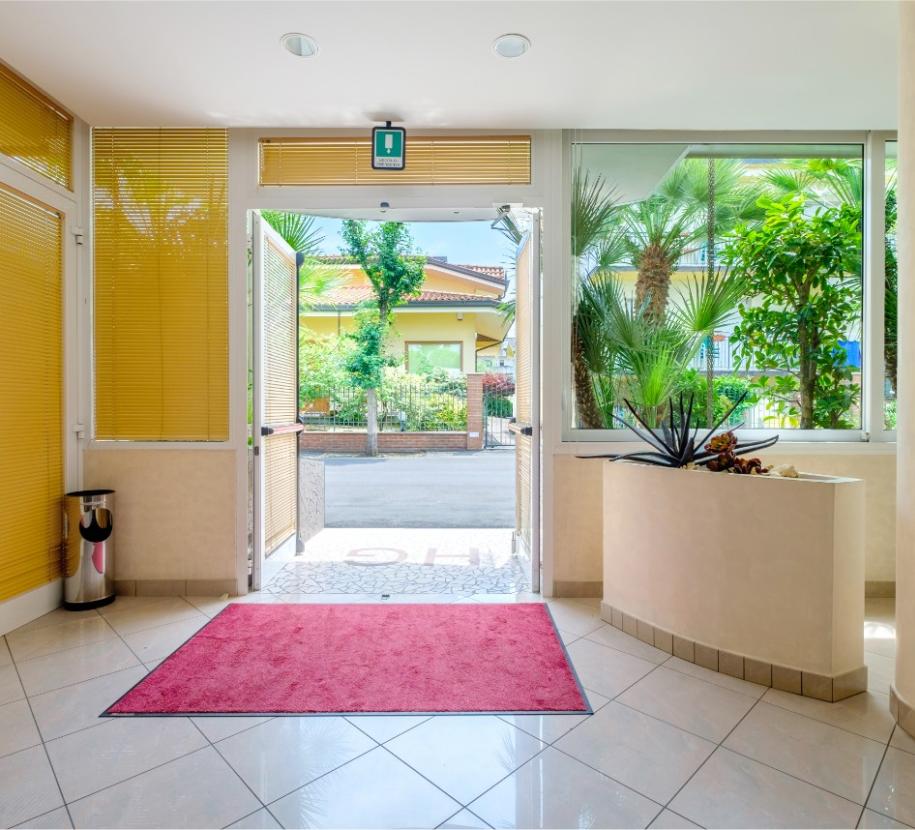 Ingresso luminoso con tappeto rosso, piante verdi e vista su giardino soleggiato.